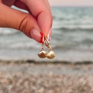 May include: Gold-coloured seashell earrings with leverback closures. The earrings are held up against a blurred background of the ocean and beach. The earrings have a textured surface and a small, dangling shell design.