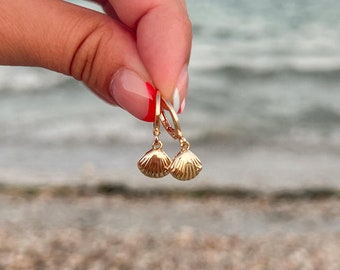 May include: Gold-colored seashell earrings with leverback closures. The earrings are held up against a blurred background of the ocean and beach. The earrings have a textured surface and a small, dangling shell design.
