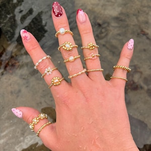 May include: A hand adorned with multiple gold rings, showcasing designs with pearls and floral motifs. The nails are painted with pink and burgundy nail polish, adding a touch of colour to the display.