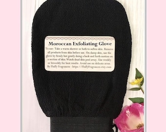 Peut inclure: Un gant exfoliant marocain noir avec une surface texturée et un bracelet élastique. Le gant porte une étiquette rectangulaire avec des instructions. Une rose rose et une fleur jaune sont en arrière-plan.