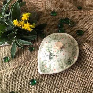 Peut inclure: Un plat en céramique blanc avec un bouchon en liège, orné d'un motif floral et du texte "Leslie Daughli+". Le plat est entouré de perles de verre vertes, de fleurs jaunes et de feuilles de sauge sur une surface en toile de jute.