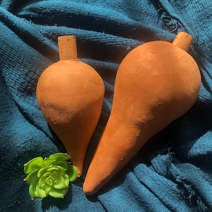 Può includere: Due bottiglie a forma di pera, color terracotta, con tappi di sughero. Una piccola succulenta verde è posta vicino alle bottiglie su un tessuto strutturato color verde acqua. Le bottiglie hanno un aspetto rustico e fatto a mano.