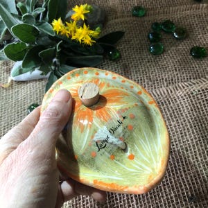 Peut inclure: Un plat en céramique peint à la main avec un bouchon en liège. Le plat présente un motif floral jaune et orange avec le texte "Lampe Divali". Le fond comprend des perles de verre vertes et du feuillage.