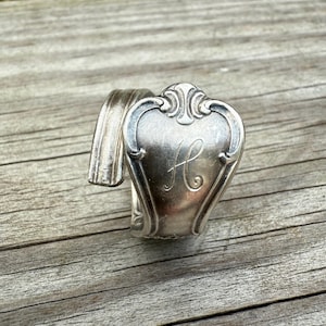 May include: A silver spoon repurposed into a bracelet. The spoon's handle is bent to form the bracelet's band, with the spoon's decorative head featuring an ornate design and the letter "H" engraved in the center.