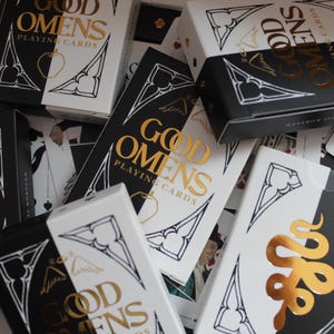 Puede incluir: Naipes con un diseño en blanco y negro. Las cajas de cartas tienen las palabras "Good Omens Playing Cards" en letras doradas. Las cajas y las cartas presentan una ilustración de serpiente dorada.