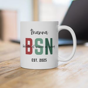 May include: White ceramic mug with the name "Brianna" and the letters "BSN" in red and green. The words "bachelor of science in nursing" are below the letters, and "EST. 2025" is at the bottom. A laptop is in the background.