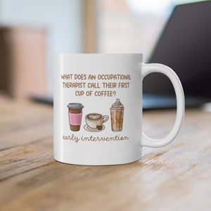 May include: White ceramic coffee mug with the text "WHAT DOES AN OCCUPATIONAL THERAPIST CALL THEIR FIRST CUP OF COFFEE?" and "early intervention". The mug features illustrations of three different coffee drinks.