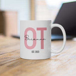 May include: White ceramic mug with the letters "OT" in pink and the name "Brianna" written in cursive underneath. The text "EST. 2023" is printed below the name.