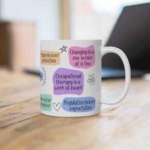 May include: White ceramic mug with colorful text and illustrations. The text reads "Progress over perfection", "Changing lives one session at a time", "Occupational therapy is a work of heart", and "Regulation before expectation".