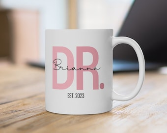 May include: White ceramic mug with a pink "DR." design and the name "Brianna" written in cursive below. The mug also has the text "EST. 2023" printed on the bottom.