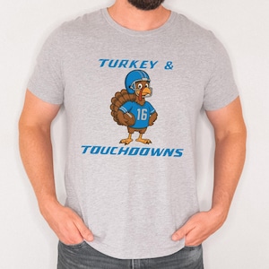 May include: A light gray t-shirt featuring a cartoon turkey wearing a football helmet and jersey with the number 16. The text "TURKEY & TOUCHDOWNS" is printed above and below the turkey illustration in blue.