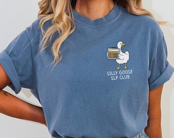 May include: A blue t-shirt with rolled-up sleeves featuring a cartoon goose holding a calculator. The text "SILLY GOOSE SLP CLUB" is printed below the goose. The shirt is made of a soft material.