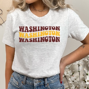 May include: A heather grey t-shirt with a retro-style graphic design featuring the word "WASHINGTON" repeated three times in a layered effect. The text is in maroon and yellow.