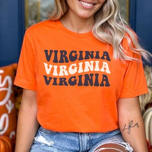 Peut inclure: T-shirt orange avec le mot "VIRGINIA" r&eacute;p&eacute;t&eacute; trois fois dans un style de police r&eacute;tro. Le texte est en noir et blanc.