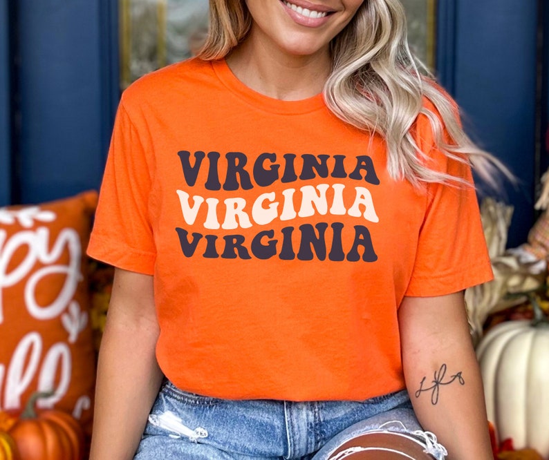 Peut inclure: T-shirt orange avec le mot "VIRGINIA" r&eacute;p&eacute;t&eacute; trois fois dans un style de police r&eacute;tro. Le texte est en noir et blanc.