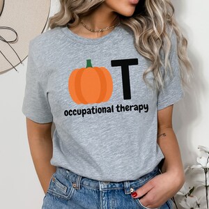 May include: A heather gray t-shirt featuring a pumpkin graphic with the letters "OT" and the words "occupational therapy" in black. The pumpkin is orange with a green stem. The shirt is made of a soft material.