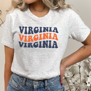 Peut inclure: Un t-shirt gris avec le mot "VIRGINIA" r&eacute;p&eacute;t&eacute; trois fois dans une police r&eacute;tro et ondul&eacute;e. Le texte est en bleu marine et orange.