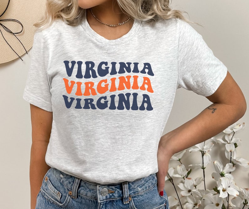 Peut inclure: Un t-shirt gris avec le mot "VIRGINIA" r&eacute;p&eacute;t&eacute; trois fois dans une police r&eacute;tro et ondul&eacute;e. Le texte est en bleu marine et orange.