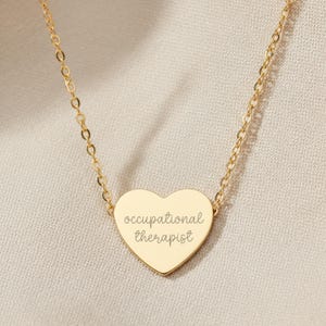 Könnte beinhalten: Eine goldfarbene Halskette mit einem herzförmigen Anhänger. Der Anhänger trägt die Inschrift "occupational therapist" in kursiver Schrift. Die zarte Kette verbindet sich mit dem Herzanhänger.
