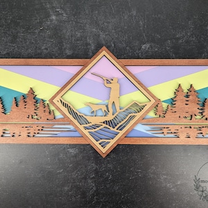 May include: Wooden layered wall art depicting a hunter and dog in a mountain scene, set against a colourful geometric background. The artwork includes laser-cut trees and a diamond-shaped central design, framed in brown wood.