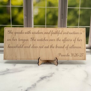 以下が含まれることがあります： 「She speaks with wisdom, and faithful instruction is on her tongue. She watches over the affairs of her household and does not eat the bread of idleness. Proverbs 31:26-27」という文章が書かれた木製の看板。筆記体で書かれている。
