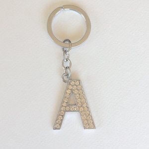 Può includere: Un portachiavi argentato con un ciondolo a forma di lettera A. La lettera A è ricoperta di strass trasparenti.