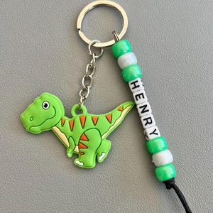 May include: A green dinosaur keyring with orange detailing and the name "HENRY" spelled out on a beaded strand. The keyring has a silver ring and chain.
