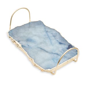 Op de afbeelding: Een rechthoekig serveerblad met een blauw en wit gemarmerde stenen plaat met een natuurlijke, ruwe rand. Het blad is voorzien van twee boogvormige, goudkleurige metalen handgrepen aan de zijkanten die contrasteren met het stenen oppervlak.