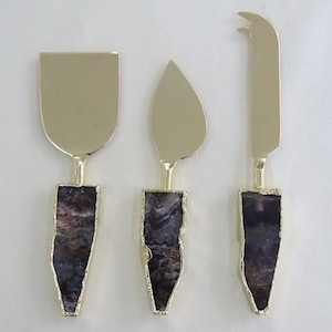 May include: Set of three gold cheese knives with agate handles. The knives have different blade shapes, including a spreader, a pointed knife, and a cheese knife.