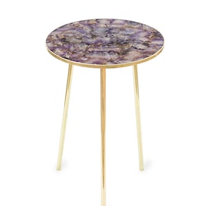 May include: A small, round side table with a tabletop made of a mosaic of purple and white stones. The tabletop is framed with a gold-colored edge. The table has three slender, gold-colored legs.
