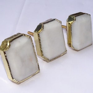 May include: Three white rectangular cabinet knobs with gold metal trim. The knobs have a slightly beveled edge and a smooth, polished finish.