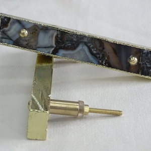 Könnte beinhalten: Goldene Schrankbeschläge mit einem rechteckigen Einlegeteil aus Achat. Der Stein hat braune, weiße und graue Muster. Die Beschläge haben eine Schraube zur Montage.