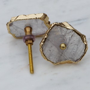 Op de afbeelding: Twee witte kwartskristal-kastknoppen met gouden metalen afwerking. De knoppen hebben een unieke, onregelmatige vorm en zijn bevestigd aan gouden metalen schroeven.