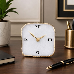 May include: A square-shaped desk clock featuring a white stone face with a natural, irregular edge finished in gold. The clock displays black Roman numerals at the twelve, three, six, and nine o'clock positions, complemented by gold-colored hands.