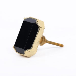 May include: A black rectangular cabinet knob with a gold-coloured, textured edge and a brass screw. The knob is angled, showing its polished black surface and the contrast with the gold trim. Suitable for adding a touch of elegance to furniture.