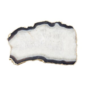 May include: A polished agate slice coaster featuring a translucent white crystalline center surrounded by concentric bands of dark charcoal and black. The irregular, organic outer edge of the stone is finished with a metallic gold-toned trim.