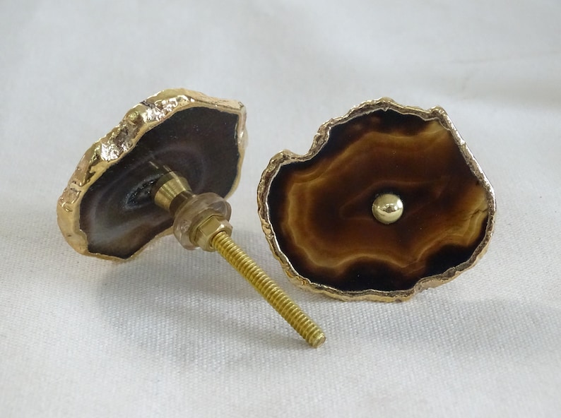 以下が含まれることがあります： 金色の縁取りが施された、2つの茶色の瑪瑙製の引き出しノブ。ノブは自然で不規則な形をしており、金色のネジで取り付けられています。