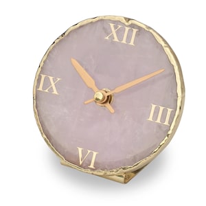 May include: A small, round clock with a light purple face and gold Roman numerals. The clock has gold hands and a gold-coloured metal frame. The clock is a decorative item.