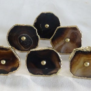 以下が含まれることがあります： 金色の縁取りが施されたアゲート製の引き出しノブ6個。各ノブは、中心に金色のつまみがあり、茶色と白のアゲートのユニークなパターンが特徴です。