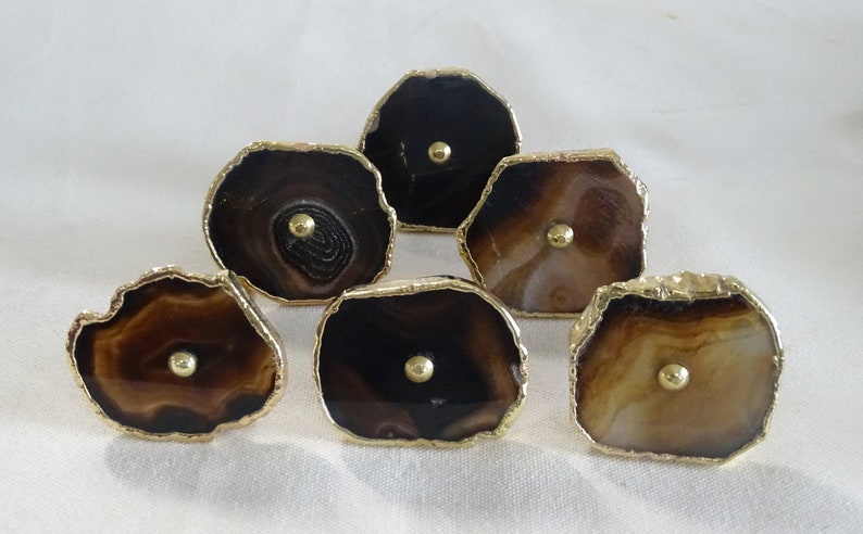 以下が含まれることがあります： 金色の縁取りが施されたアゲート製の引き出しノブ6個。各ノブは、中心に金色のつまみがあり、茶色と白のアゲートのユニークなパターンが特徴です。