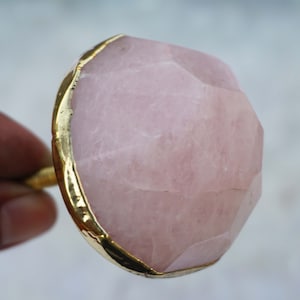 Pode incluir: Um botão de quartzo rosa rosa com uma borda dourada. O botão é facetado e tem um acabamento polido.