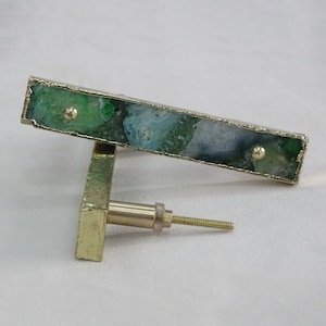 Puede incluir: Un tirador rectangular para armario con un centro de piedra de ágata verde y blanca, enmarcado en metal dorado. El tirador tiene una base y un tornillo dorados para la instalación. Herrajes decorativos para muebles.