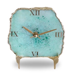 Puede incluir: Un reloj de ágata azul turquesa con números romanos en un soporte dorado. El reloj tiene una aguja de hora y una aguja de minutos doradas.
