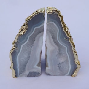 May include: Two agate bookends with a gold-coloured trim. The bookends are a light grey colour with white and darker grey bands. The edges are trimmed with a textured gold colour. The bookends are cut in a vertical orientation.