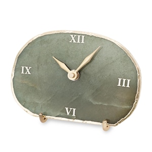 Puede incluir: Un reloj de escritorio ovalado con una esfera de piedra verde salvia y un borde natural de acabado dorado. El reloj muestra números romanos blancos en las posiciones de las doce, tres, seis y nueve, junto con dos manecillas de color dorado.