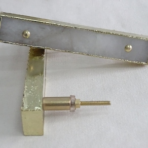 Puede incluir: Un tirador rectangular para armario con marco dorado y centro gris claro. Los bordes del marco tienen un aspecto texturizado. Dos pequeños remaches dorados aseguran la pieza central. El tirador está fijado a una base rectangular dorada.