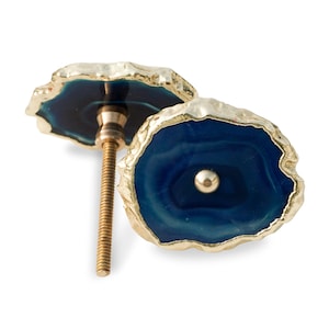 Può includere: Due pomelli decorativi per mobili. Ogni pomello presenta un centro blu scuro, in stile agata, con un bordo irregolare color oro. I pomelli hanno una vite color oro e un piccolo accento centrale color oro.