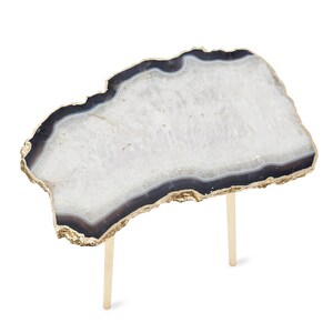 May include: A side table featuring an irregular, slab-like top made of white and dark gray agate stone with a natural, rough-hewn edge finished in metallic gold. The tabletop is supported by two slender, cylindrical legs in a matching gold tone.