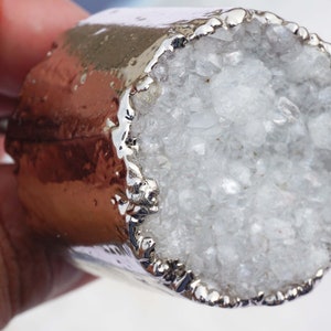 Könnte beinhalten: Ein silberner Metallknopf mit einem weißen Kristallgeode, der oben eingebettet ist. Der Geode ist von einem silbernen Metallrand umgeben.