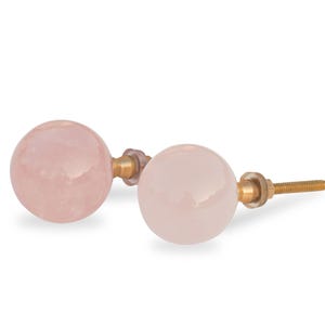 May include: Two pink round knobs with gold metal hardware. The knobs are made of rose quartz and have a smooth, polished finish.